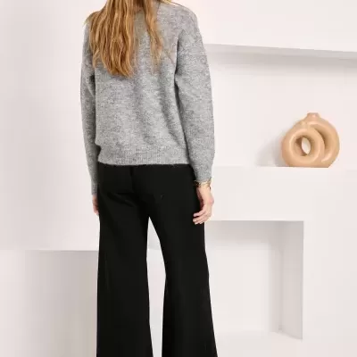 Mulher de costas com camisola cinzenta e calças pretas largas em fundo branco com objetos decorativos