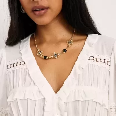 Colar dourado decorativo com pedras pretas e pérolas, em modelo com blusa branca rendada.