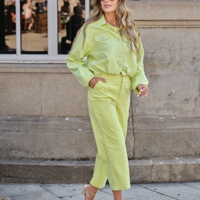 Mulher de cabelo loiro ondulado com macacão verde-limão e sapatos dourados, numa rua com parede de pedra.