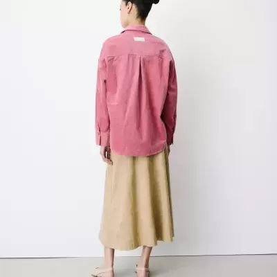 Mulher com camisa rosa de veludo cotelê, saia beje longa e sandálias bejes em fundo branco.