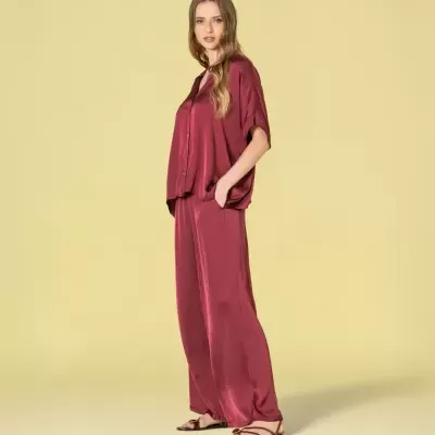 Modelo veste conjunto vermelho escuro acetinado com camisa e calças largas e sapatos rasteiros castanhos