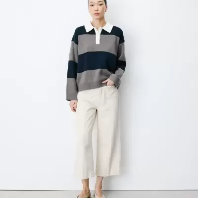 Mulher com camisola de malha com riscas cinzentas e azuis, calças creme largas e sapatos rasos beges.
