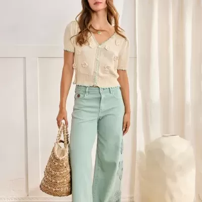 Mulher com camisola creme e calças de ganga azuis claras com aplicação de flores, mala de palha e sandálias, em ambiente interior com cortinas brancas.