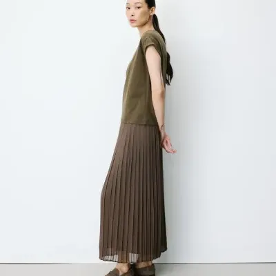 Mulher com t-shirt verde-oliva, saia plissada castanha e mocassins castanhos junto a parede branca.