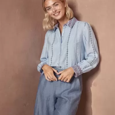 Mulher com camisa azul clara bordada e calças azul cinzento em fundo bege