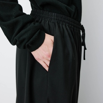 Camisola preta de malha com calças pretas de tecido liso e cinto elástico com cordão com mão no bolso.
