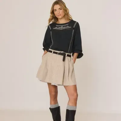 Mulher vestindo camisola preta com bordados, saia bege com cinto, meias cinzentas e botas pretas em estúdio branco.