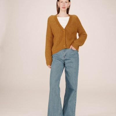 Mulher com camisola castanha, jeans largos azuis e sapatos pretos sobre fundo bege