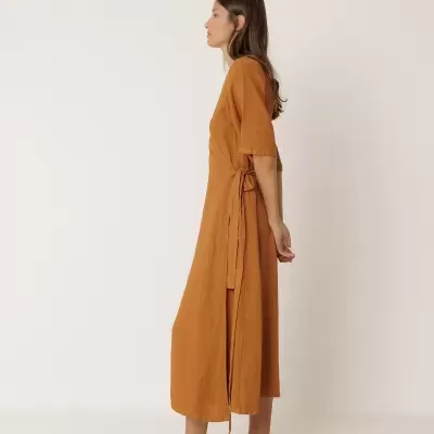 Vestido comprido de linho castanho com cinto lateral ajustável, mangas curtas, e sapatos baixos cinzentos, mulher vista de perfil.