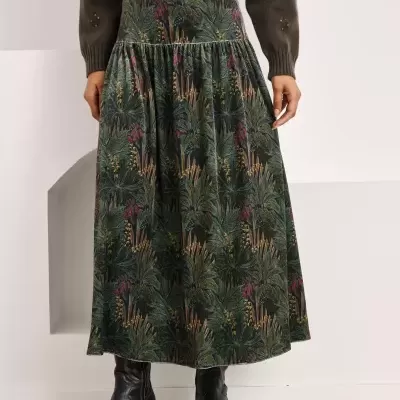 Saia comprida com padrão de folhagem verde e suéter castanho com detalhes em renda e botões, botas pretas com bordado.