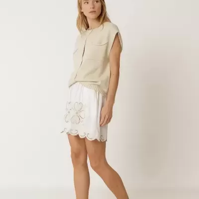 Mulher com camisa de malha bege sem mangas, saia branca bordada e chinelos escuros