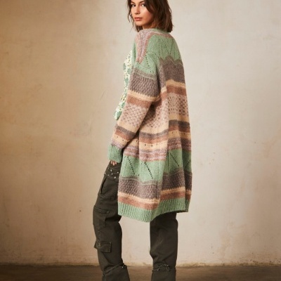 Mulher a usar casaco comprido de malha com riscas coloridas, calças cargo verdes e botas pretas em sala com parede bege.