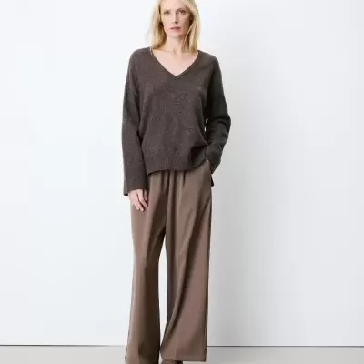 Mulher loira com camisola castanha, calças largas castanhas e sapatos castanhos em fundo branco