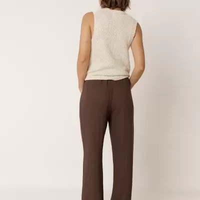 Pessoa de costas com calças largas castanho-escuras, colete creme e sandálias castanhas.