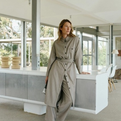 Mulher vestindo conjunto cinzento às riscas com casaco comprido e calças largas, sapatos brancos, ambiente interior moderno e luminoso