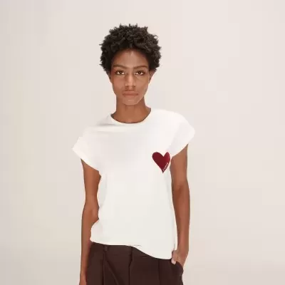 T-shirt branca com coração vermelho bordado e calças castanhas.