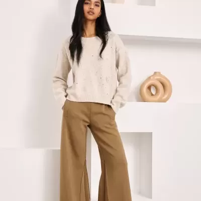 Mulher com camisola bege clara e calças largas castanho claro em ambiente minimalista.