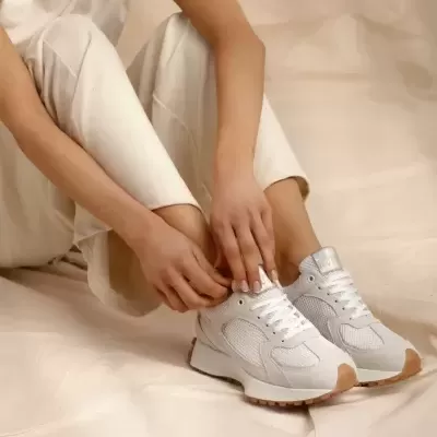 Ténis desportivos brancos com detalhes cinza e sola castanha usados por pessoa com calças claras sentada.