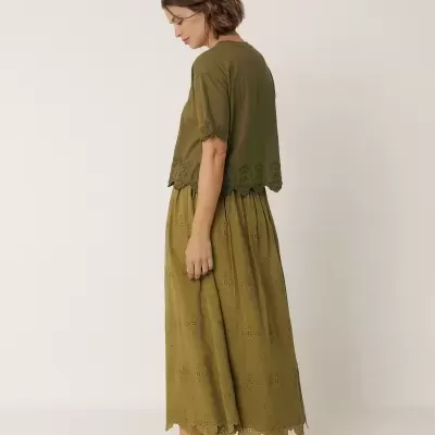 Mulher de perfil com camisa e saia verde-oliva com bordados e sandálias castanhas