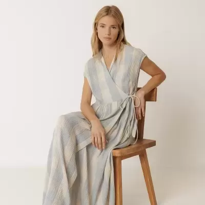 Mulher sentada numa cadeira de madeira com vestido comprido às riscas azul claro e bege e sapatos azuis claros.