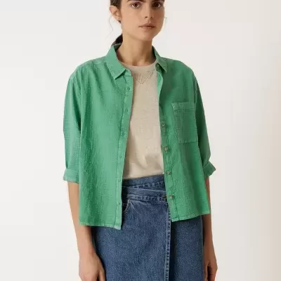 Mulher com camisa verde, camiseta bege e saia de ganga azul em fundo branco