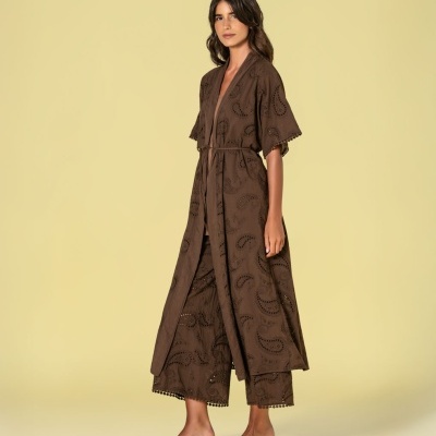 Modelo feminino veste conjunto castanho com vestido longo e calças largas, com padrão bordado paisley e pompons nas bainhas, e sandálias pretas de salto médio