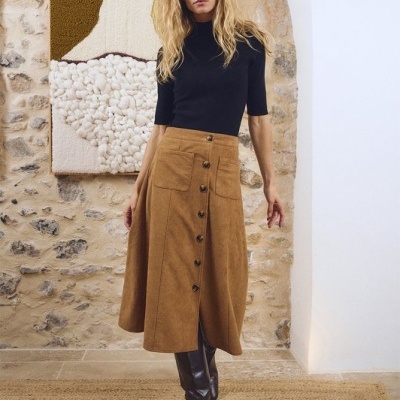 Mulher com saia castanha de botões, camisola preta e botas pretas, parede de pedra e tapete castanho no interior