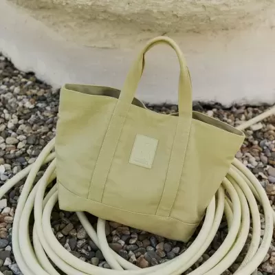 Bolsa de tecido bege com alças curtas e logo, sobre mangueira enrolada em seixos