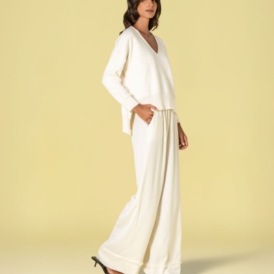 Mulher vestindo conjunto branco com camisola de manga comprida e calças largas, sandálias pretas, fundo amarelo