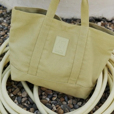 Mala tote de tecido verde-oliva com etiqueta e alças, sobre mangueira e pedras