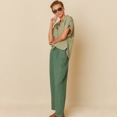 Pessoa com calças verdes largas, camisa verde claro quadriculada, óculos escuros castanhos, em fundo bege.