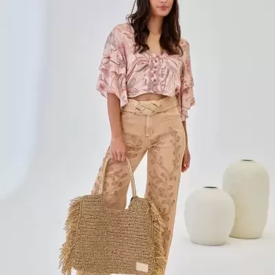 Mulher com blusa rosa floral, calças bege bordadas, sandálias plataforma e mala de ráfia
