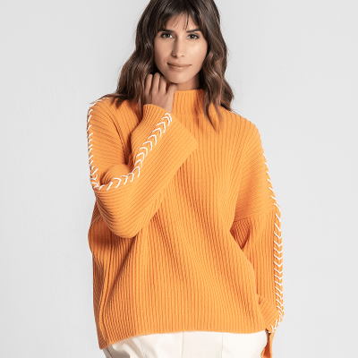 Mulher com camisola laranja de malha grossa e calças brancas em fundo branco