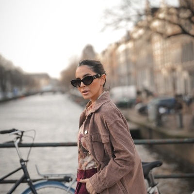 Mulher com casaco canelado castanho e óculos de sol junto a bicicletas junto a um canal