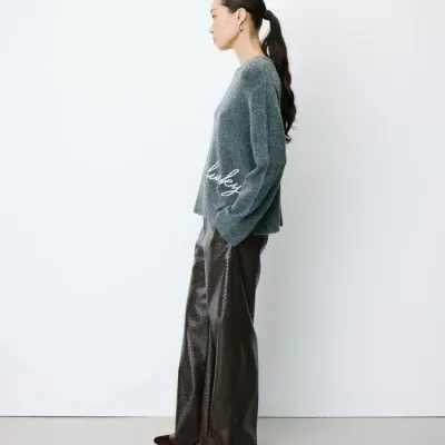 Mulher de perfil com camisola cinza e calças largas marrons com textura