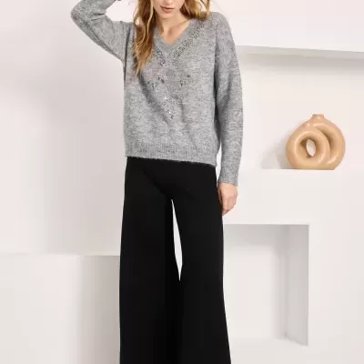 Mulher com casaco cinza de malha e calças largas pretas em ambiente interior branco com prateleiras decorativas.
