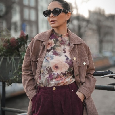 Mulher com casaco castanho claro de veludo cotelê e blusa floral, calças vinho, óculos escuros, junto a bicicleta na rua