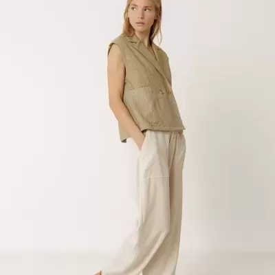 Mulher com colete às riscas verdes e bege, calças largas bege claras e mocassins castanhos claros numa superfície branca