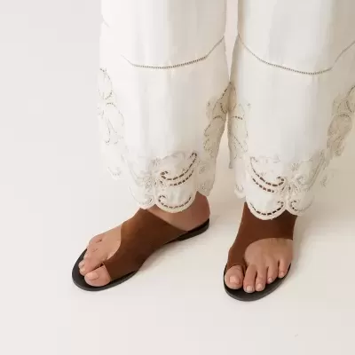 Sandálias rasas castanhas com sola preta num fundo branco e pernas com calças brancas bordadas