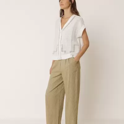 Mulher com blusa branca bordada, calças bege riscas e mocassins brancos num fundo creme