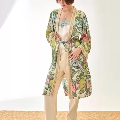 Mulher com robe estampado verde, rosa e creme, camisola branca com renda, calças claras e sandálias de plataforma em chão de tapete de palha