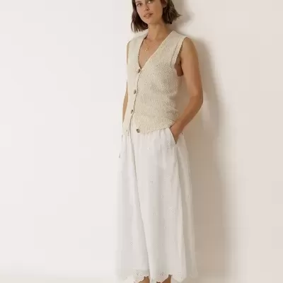 Mulher com colete de malha cru, saia branca bordada e sapatos rasos bege