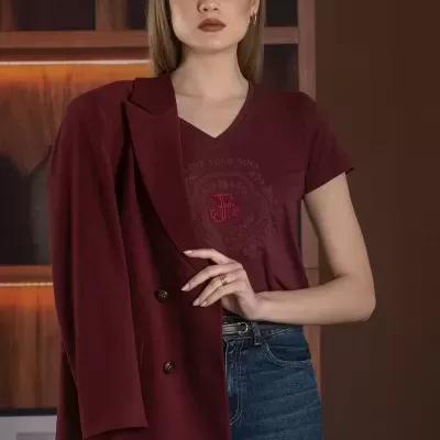 Mulher com casaco bordeaux e camisa bordeaux com texto, jeans azuis, em interior com prateleiras de madeira