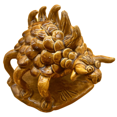 Escultura cerâmica castanha de criatura fantástica com escamas e dentes, sobre base retangular.