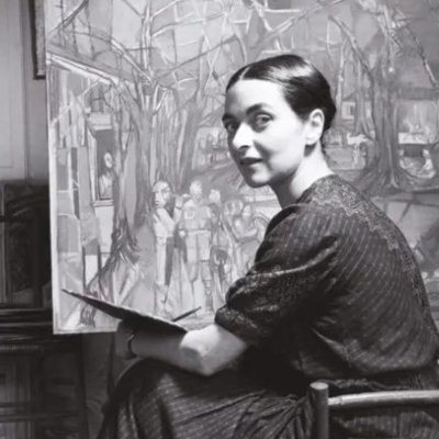 Mulher sentada a pintar junto a uma tela com pintura colorida