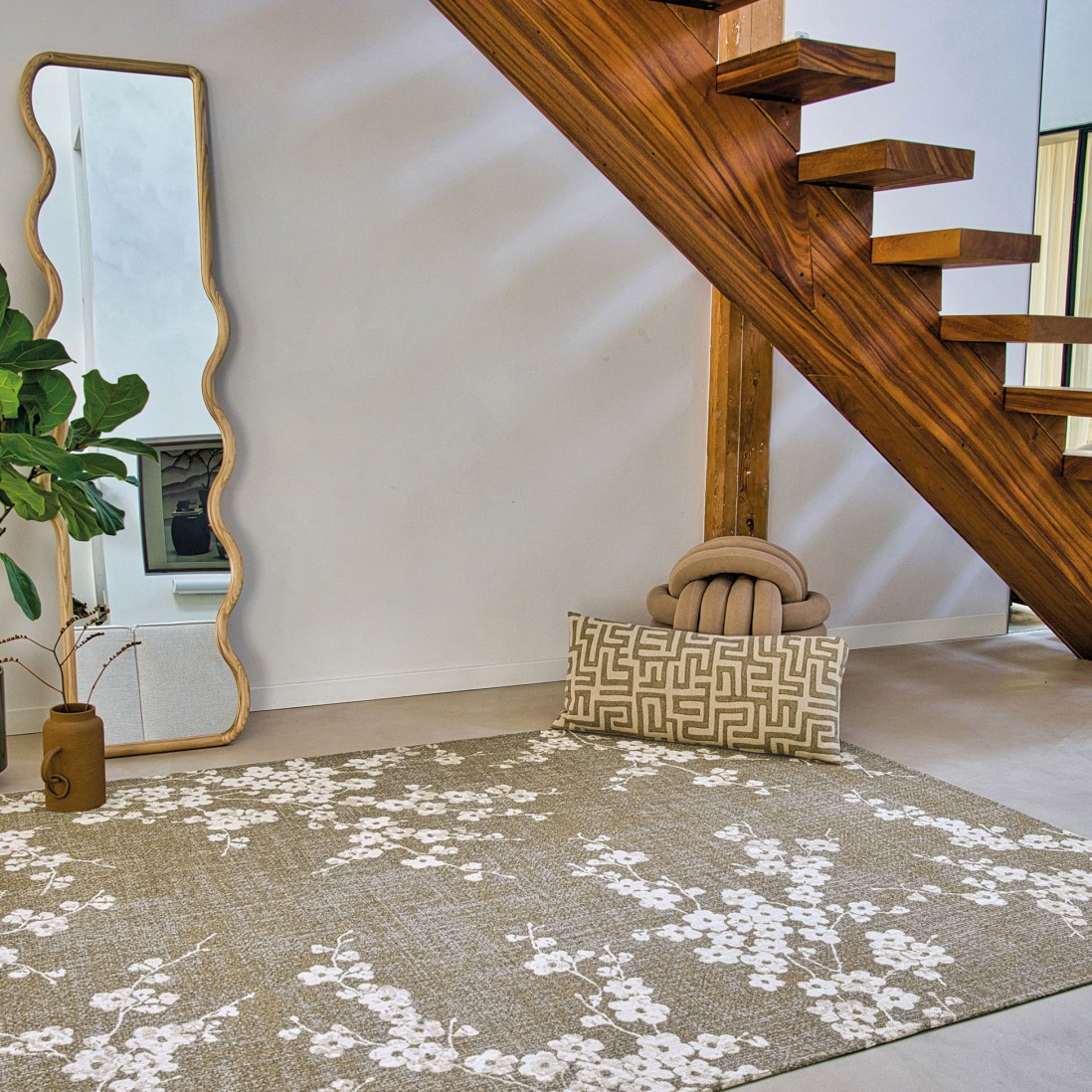 Tapete Almond Chenille Tapete bege com flores brancas debaixo de escada de madeira, espelho, vaso com plantas e almofada decorativa.
