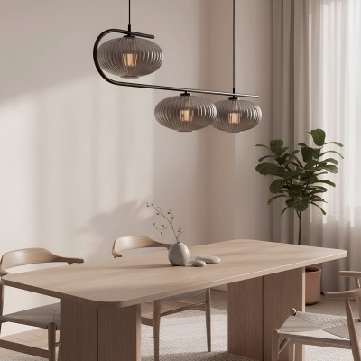 Mesa de madeira clara com quatro cadeiras, candeeiro de teto com três abajures cinzentos, vaso e pedras decorativas, planta verde e cortinas brancas.