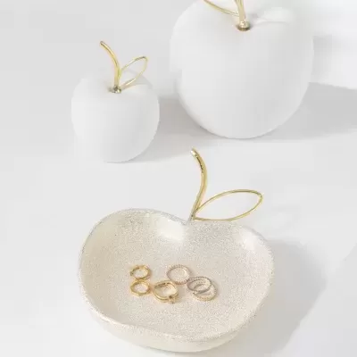 Três maçãs decorativas, uma tigela dourada com anéis metálicos em cima de superfície branca