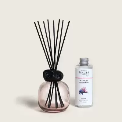 Difusor de aroma rosa com pedra preta e varetas pretas ao lado de garrafa plástica transparente de recarga com rótulo branco sobre fundo bege.