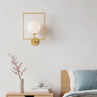 Candeeiro de parede dourado com globo branco acima de mesa de cabeceira em madeira clara com vaso branco e flores cor-de-rosa